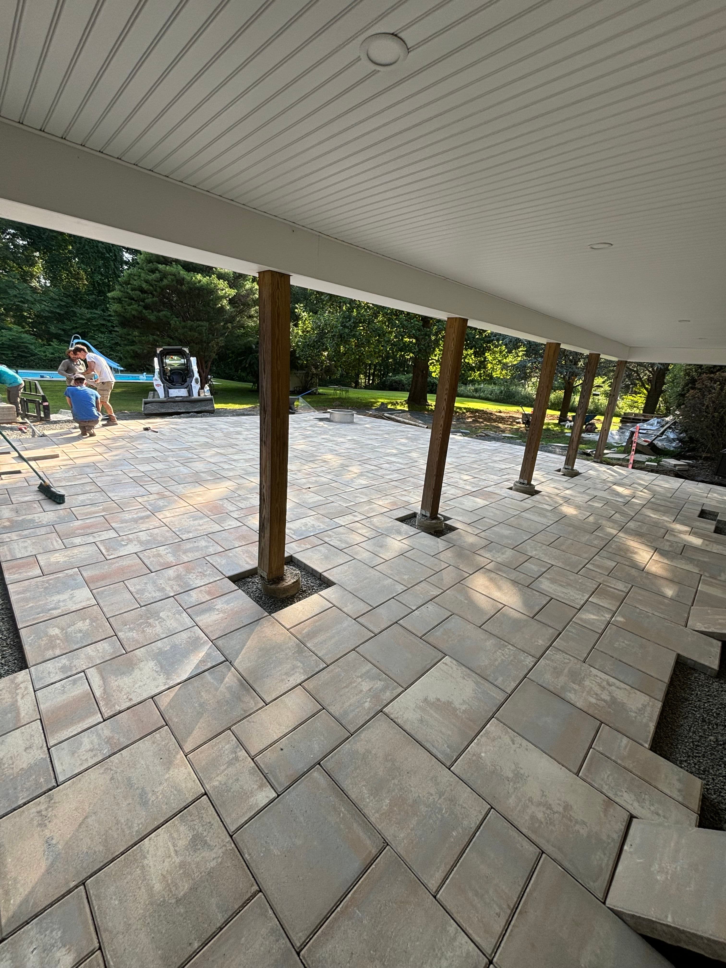 Paved outdoor patio with new tiles, supported by wooden columns, in a lush green setting.