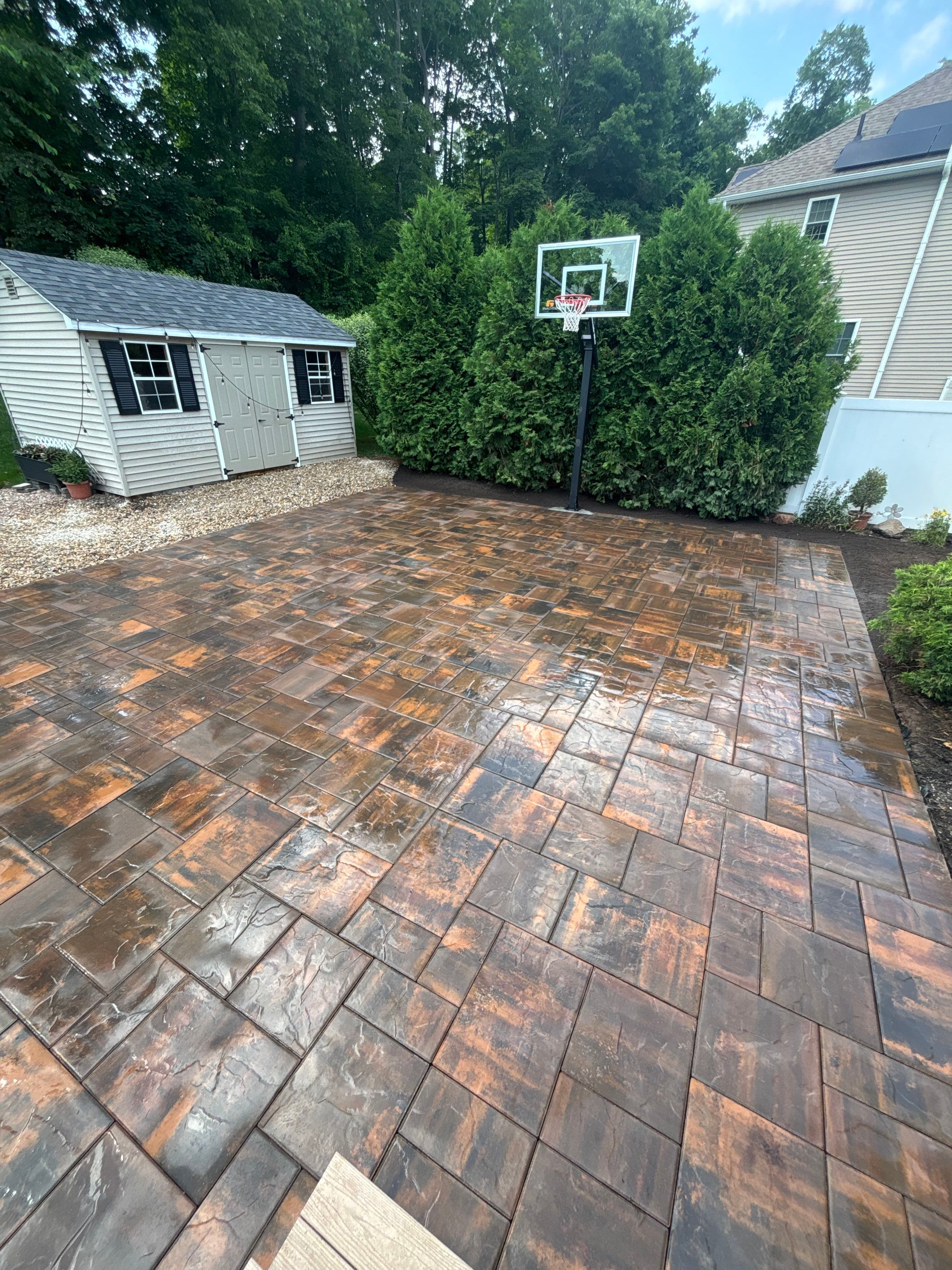 Stamped concrete basketball court in a backyard with a storage shed and greenery.