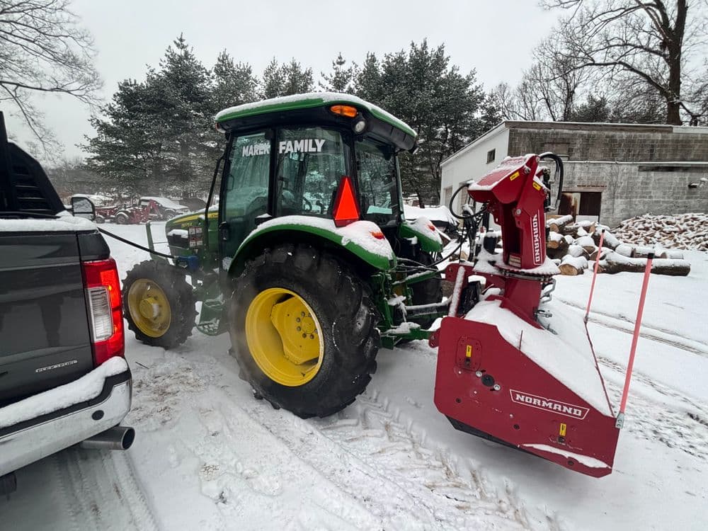 Green tractor with snowplow attachment in snowy landscape, parked beside a vehicle.