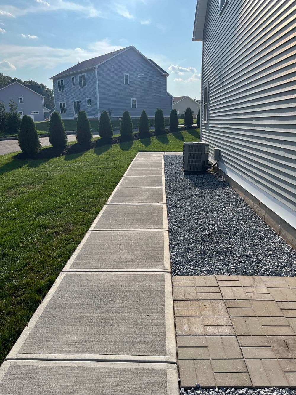 Concrete walkway alongside a house with shrubs, gravel area, and clear blue sky.
