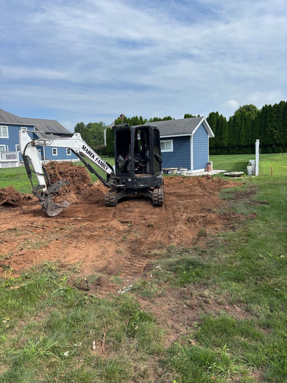 Excavator digging a yard near a blue house under a cloudy sky, with soil and grass visible.