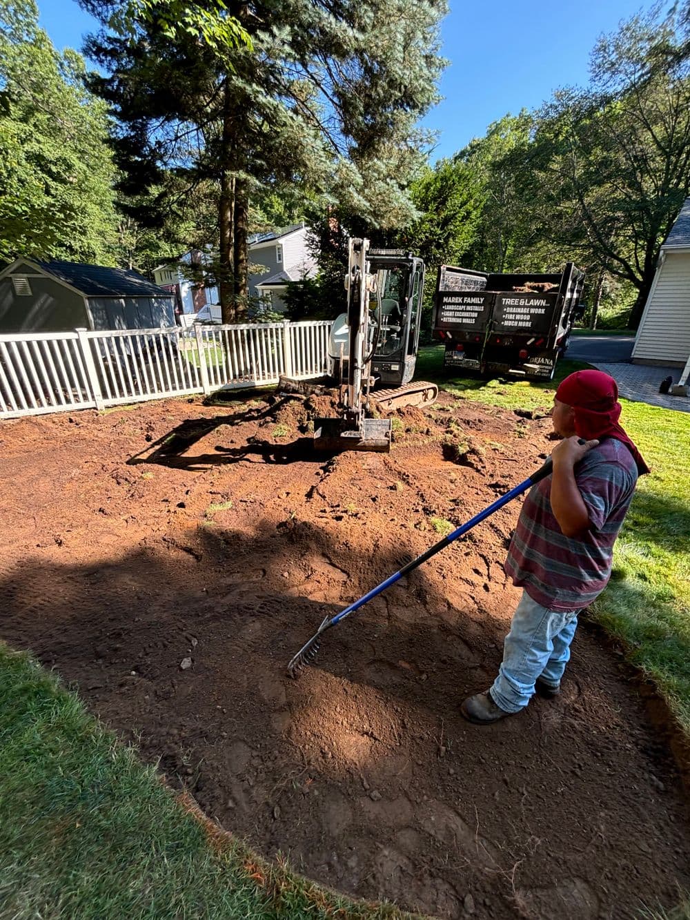 Child raking soil in a backyard, with excavation equipment in the background and trees around.