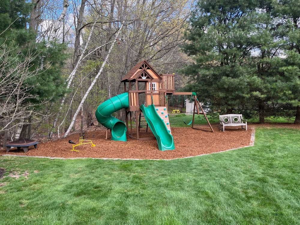 Wooden playset with slide, swing, and climbing features in a green backyard setting.