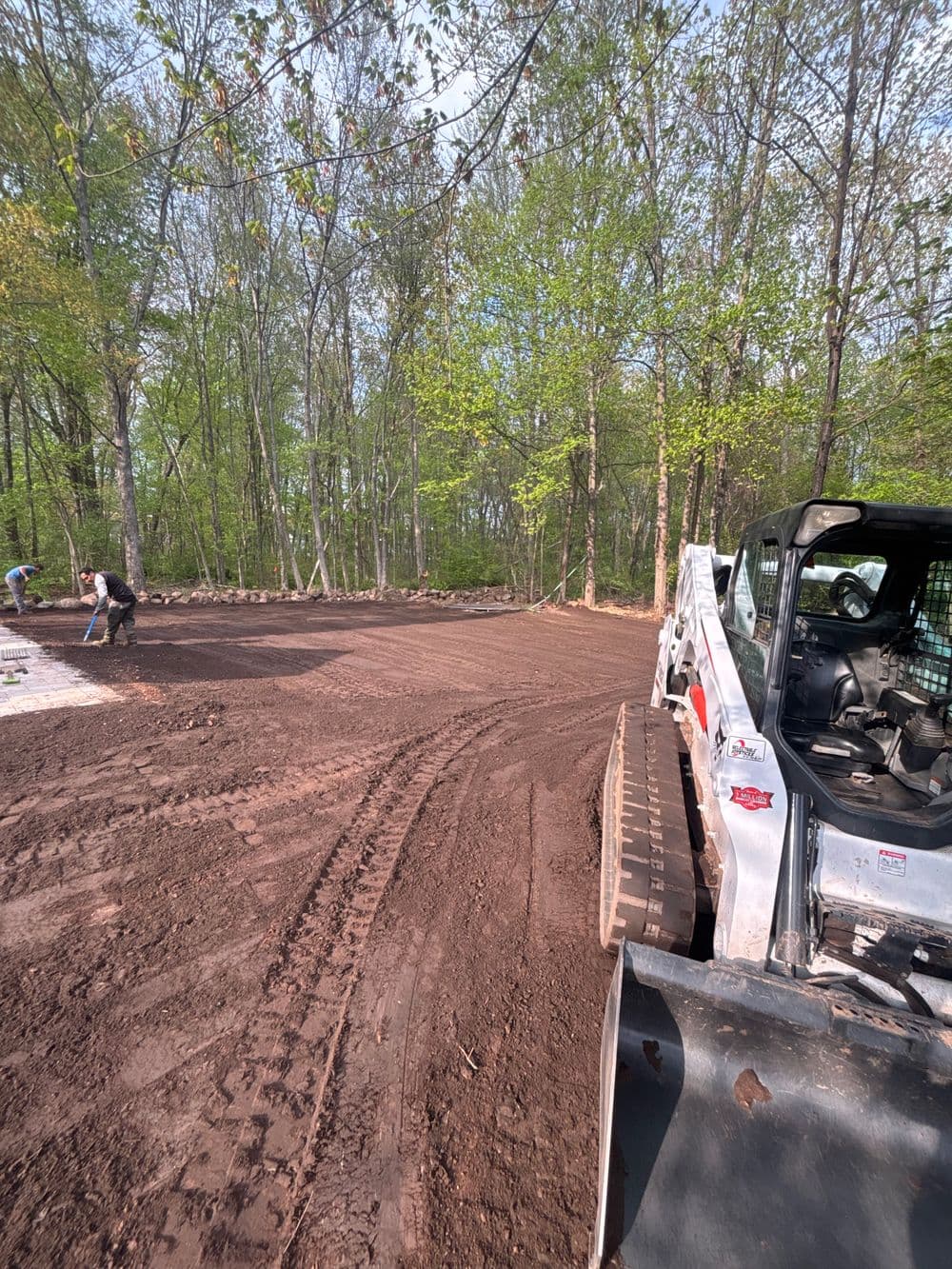 Bobcat equipment clearing land for landscaping in a wooded area with workers in the background.