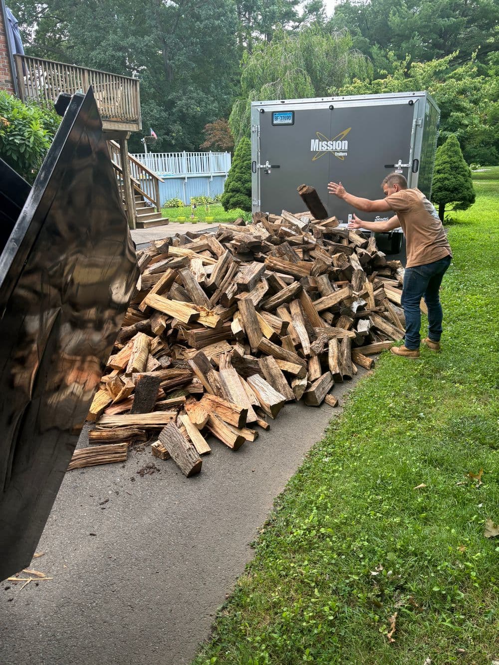 Person stacking firewood next to a delivery trailer on a grassy yard.