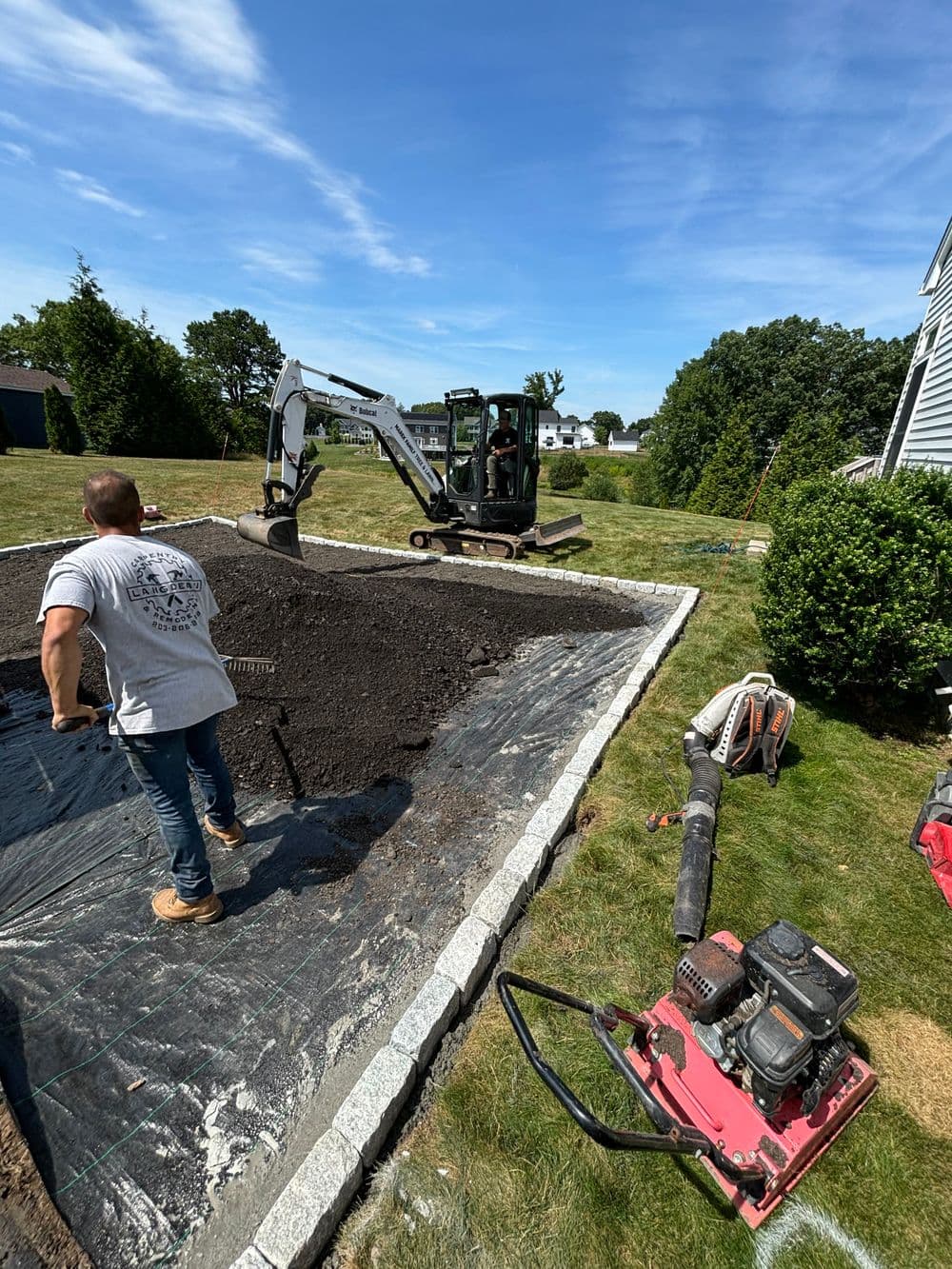 Excavator and worker preparing landscaping area with equipment and grass in background.