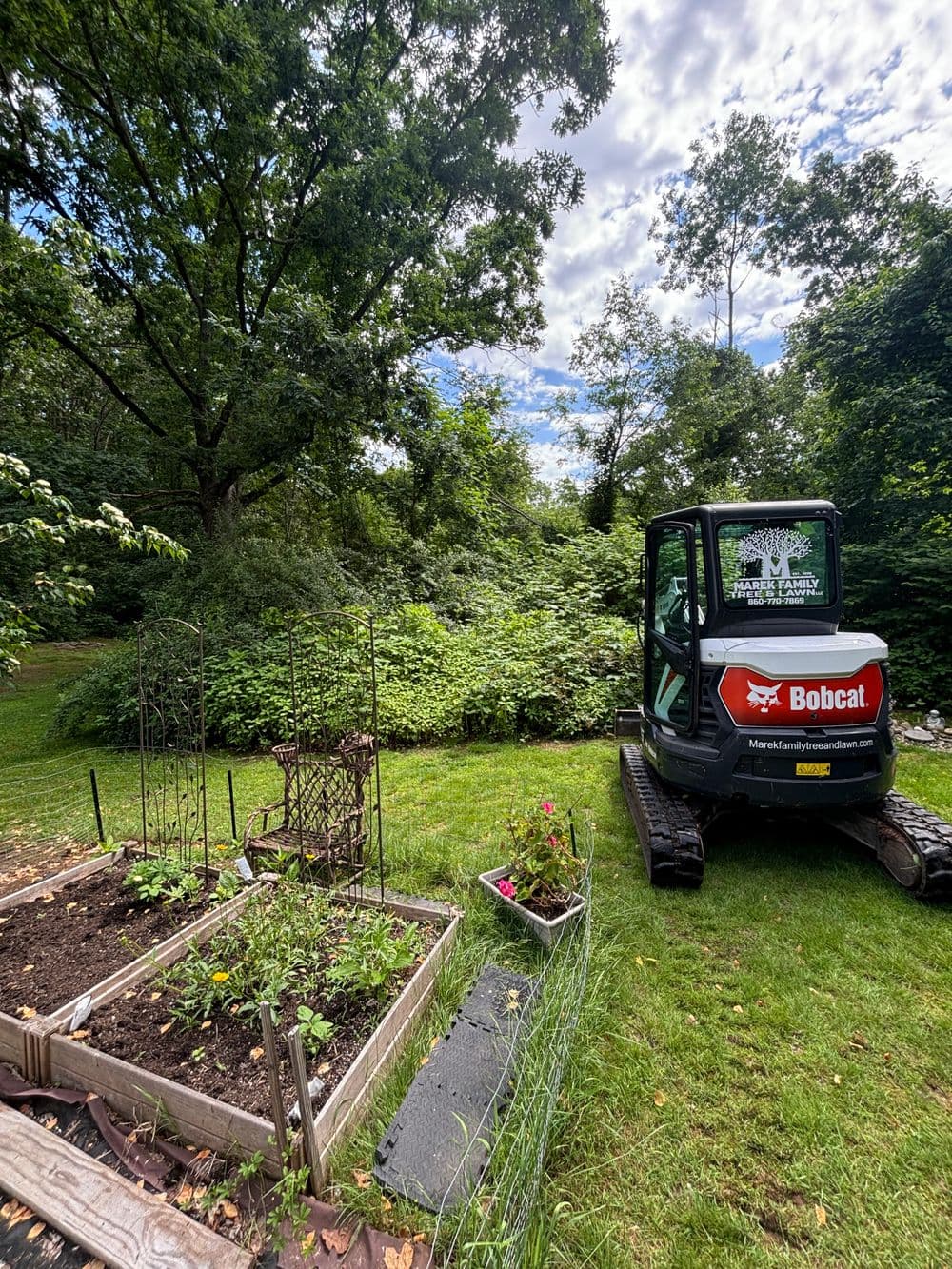 Bobcat machine next to garden beds in a landscaped outdoor setting, with trees and clouds overhead.