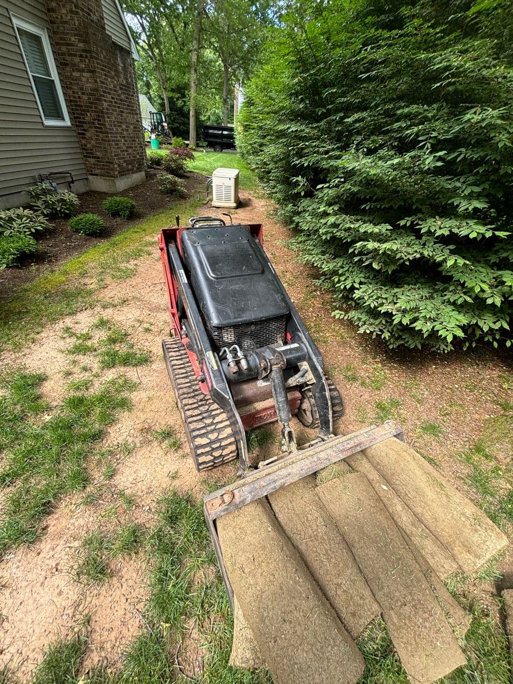 Small tracked skid steer loader on a grassy path near a home, surrounded by bushes.