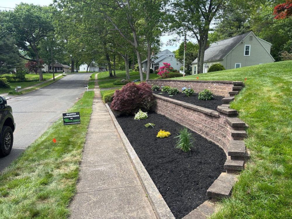 Landscaped garden with flowers and shrubs along a brick retaining wall on a residential street.