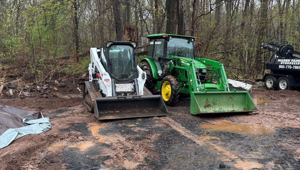 Two heavy machinery vehicles, a Bobcat skid steer and a John Deere tractor, on a construction site.