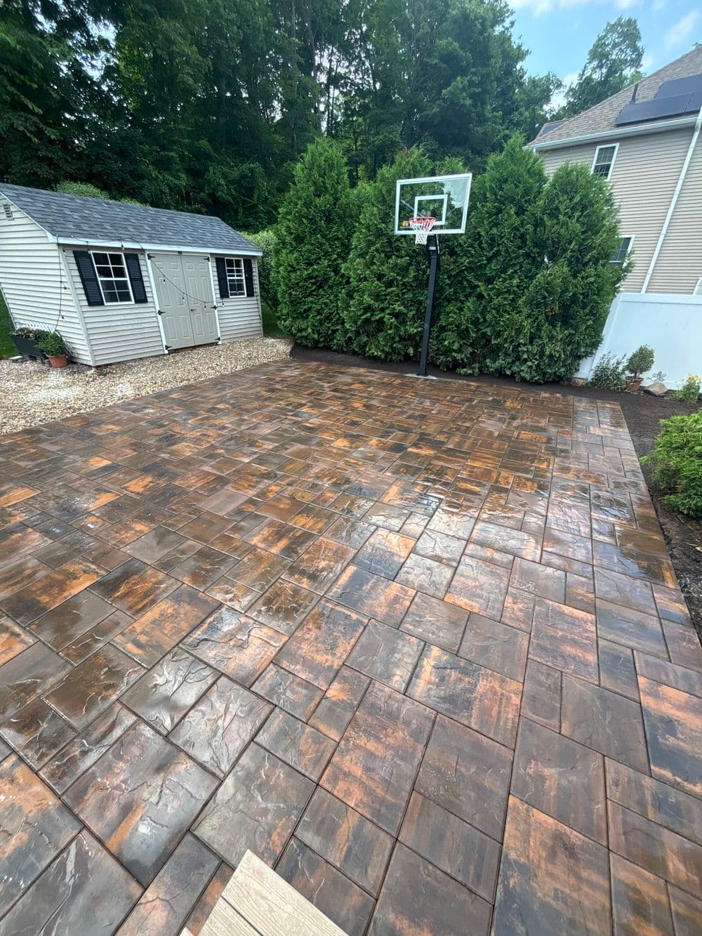 Basketball court paver patio with shed and greenery in background.